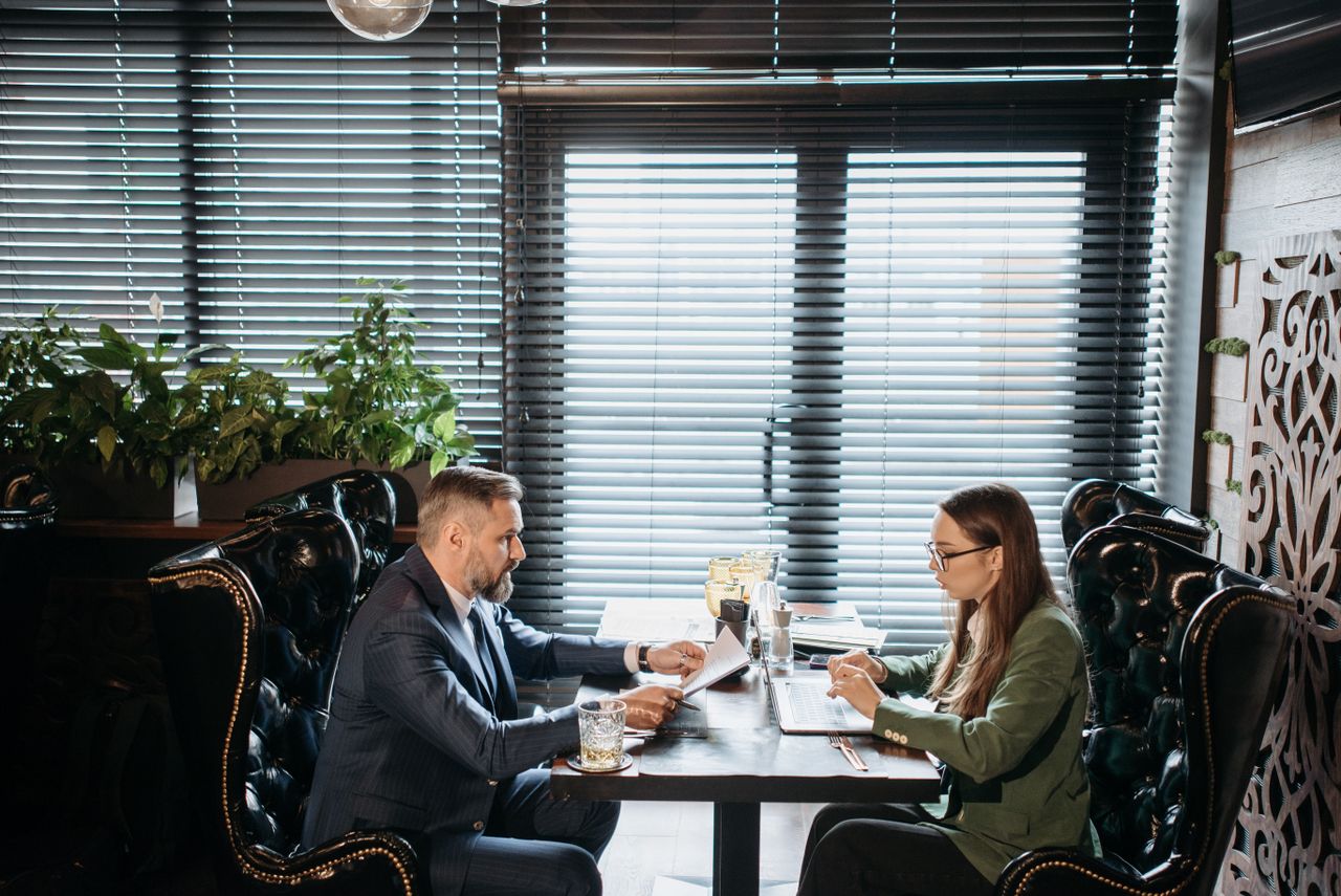 A man and woman in business attire discussing work over drinks in a stylish restaurant.